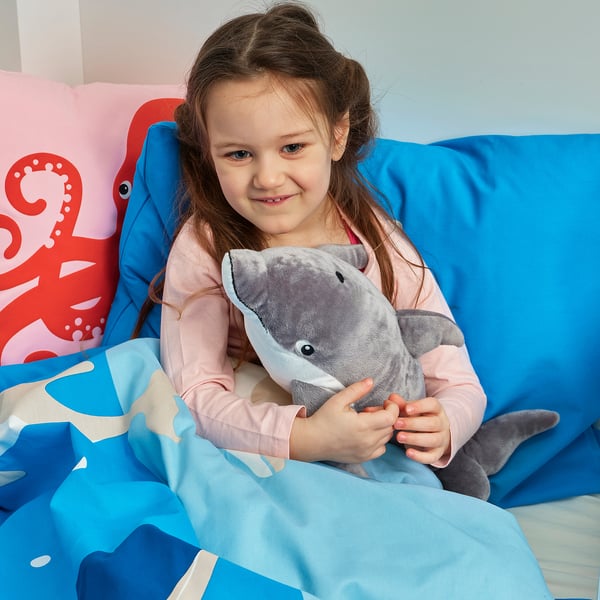 Unha rapaza abraza un xoguete de peluche de golfiño gris nunha cama azul. Ela en pixama rosa baixo unha manta azul. Unha almofada de polbo vermello está detrás dela.