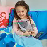 Unha rapaza abraza un xoguete de peluche de golfiño gris nunha cama azul. Ela en pixama rosa baixo unha manta azul. Unha almofada de polbo vermello está detrás dela.
