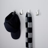 Catro ganchos metálicos de parede conteñen unha gorra negra e unha bufanda de raias, organizando ordenadamente os accesorios nun corredor.
