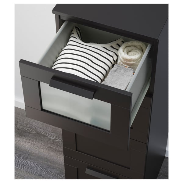 Cofre negro con caixón superior de vidro xeado, que sostén camisa e xersei raiados, caixóns de correr lisos, necesita ancoraxe na parede.