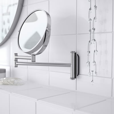 Espello de aceiro inoxidable montado con lado de lupa na parede de azulexos, reflicte a configuración limpa do baño.