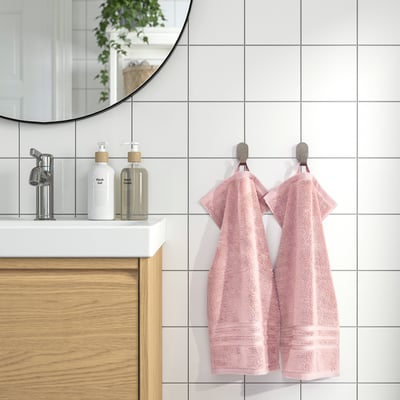 As toallas rosas BROKGLIM colgan ordenadamente en ganchos cromados xunto a un lavabo moderno con mesa branca, espello e armario de madeira. Suave, absorbente e orgánico.