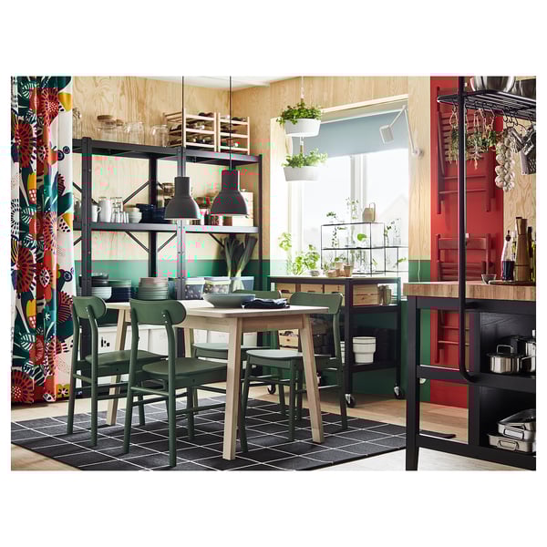 Cociña luminosa con cadeiras verdes, mesa de madeira e estantes metálicos con pratos, plantas e potas. Cortinas de cores á esquerda.