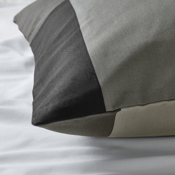 Primeiro plano da funda de almofada BRUNKRISSLA suave e de alta calidade. Tecido gris escuro cun brillo sutil, descansando sobre roupa de cama clara.
