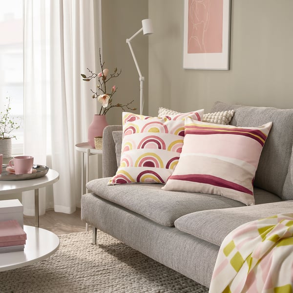 Sala de estar acolledora con sofá gris, coxíns BRUNTÅG rosa e beige, mesa lateral con plantas e cunca, lámpada cálida e manta con acento verde.