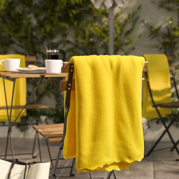Manta amarelo brillante DAGGKÅPOR, suave e reciclada, drapada sobre cadeira, máis grande que as cuncas da mesa próxima.