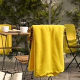 Manta amarelo brillante DAGGKÅPOR, suave e reciclada, drapada sobre cadeira, máis grande que as cuncas da mesa próxima.