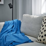 Un acolledor lanzamento azul DAGGKÅPOR, que cobre principalmente un sofá gris claro, con almofadas de puntos negros e unha lámpada preto.
