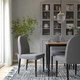 Zona de comedor moderna con cadeiras e mesa grises DANDERYD, elegantes pernas de metal negro, colocadas sobre alfombra en zigzag. Armario de vidro detrás, verde, lámpada colgante.
