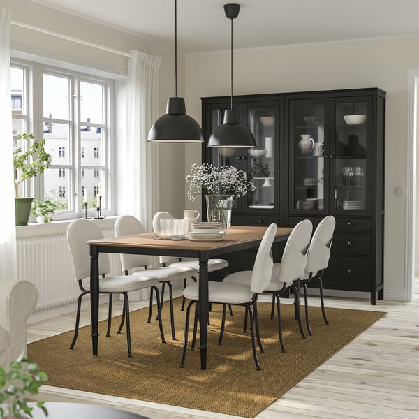 Comedor negro de 6 asentos con mesa de madeira e cadeiras brancas. Mesa con pratos, flores e lámpadas. Gran armario negro, alfombra beige.