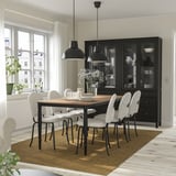 Comedor negro de 6 asentos con mesa de madeira e cadeiras brancas. Mesa con pratos, flores e lámpadas. Gran armario negro, alfombra beige.