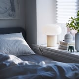 Dormitorio acolledor con lámpada branca DEJSA, roupa de cama de seda, cabeceira gris, arte azul e plantas en vaso.