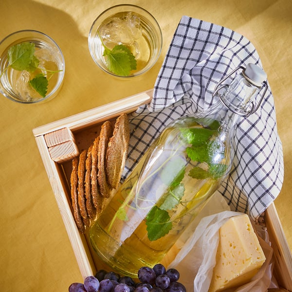 Caixa de madeira con xerra de bebida de flores de sabugueiro, queixo, uvas, vasos e pano.