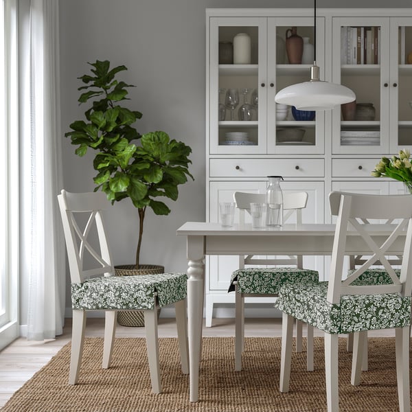 Zona de comedor moderna con mobles brancos. Dúas cadeiras florais, pano de mesa verde e planta grande. Armario branco con cristalería.