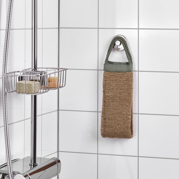 Un esfoliante corporal de xute colga nun gancho nun baño, con mangos fáciles de agarrar. Preto, un caddy de ducha cromado contén obxectos.