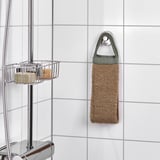 Un esfoliante corporal de xute colga nun gancho nun baño, con mangos fáciles de agarrar. Preto, un caddy de ducha cromado contén obxectos.