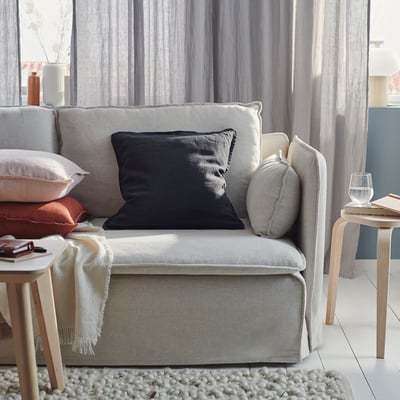Un acolledor sofá gris con almofadas negras, gris e pexegos, un lanzamento de crema, mesas laterales de madeira e un vaso de auga. Cortinas gris claras no fondo.