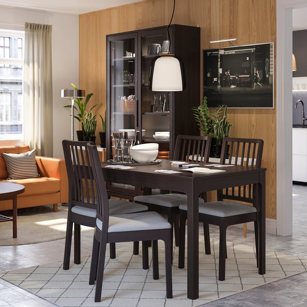 Conxunto de comedor moderno con mesa de madeira escura, cadeiras grises, armario de vidro e plantas. Espazo de estar luminoso ao fondo.