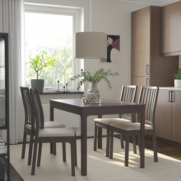 Conxunto de comedor moderno con mesa de madeira escura e cadeiras beige. Grandes fientrenas, arte, plantas.