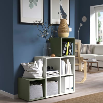 Sala de estar moderna con estante de almacenamento verde EKET que mostra libros, vasos e almofadas contra a parede gris-azul.