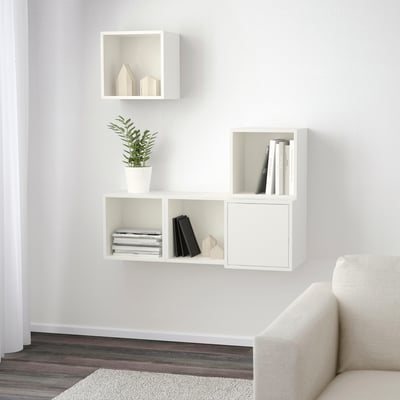 Unidade de estante de parede branca minimalista. Tres compartimentos conteñen libros, unha planta e decoración. Solución de almacenamento eficiente.