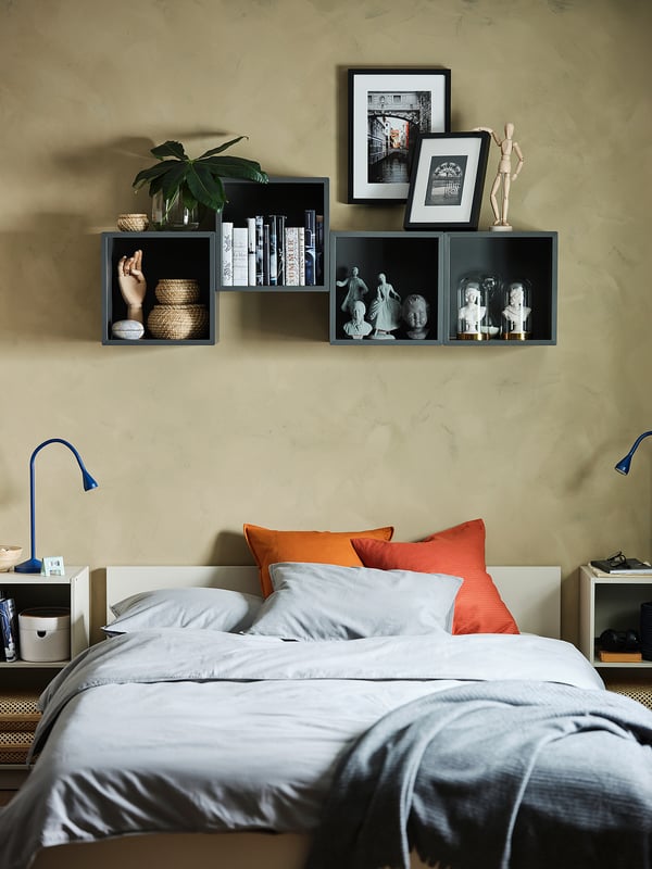 Dormitorio moderno con paredes beige, cama gris e laranxa, estante gris escuro EKET con libros e decoración.