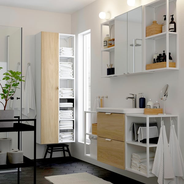 Baño moderno con armarios de madeira clara, espello, lavabo e armario de liño alto que sostén toallas.