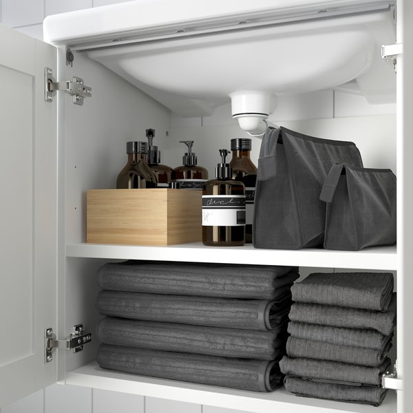 Armario de baño branco con toallas e artigos de baño. O estante superior contén botellas, bolsa e caixa de madeira.