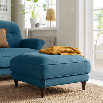 Pé azul brillante ESSEBODA con pernas de madeira, manta de mostaza dobrada na parte superior, acolledor ambiente de sala de estar.