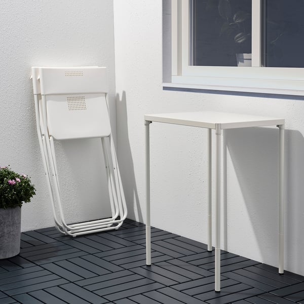 Unha cadeira plegable de metal branco apoiase contra unha parede branca xunto a unha pequena mesa branca nunha cuberta de madeira.