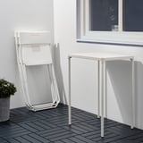 Unha cadeira plegable de metal branco apoiase contra unha parede branca xunto a unha pequena mesa branca nunha cuberta de madeira.