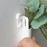 Unha man pega unha planta branca a unha parede gris, sostendo unha planta artificial verde.