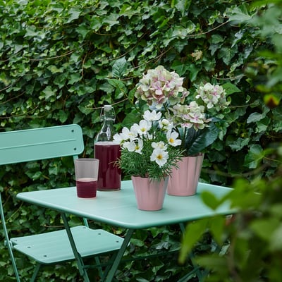 Mesa e cadeiras verdes con plantas, flores e cristalería rosas contra a vexetación.