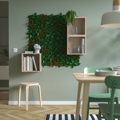 Comedor moderno con parede verde de plantas FEJKA, mesa de madeira clara, cadeiras verdes e taburete de madeira. Tons de terra e alfombra de raias.