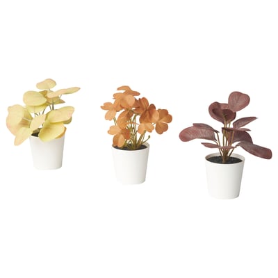 Tres plantas artificiais en vasos en recipientes brancos, de diferentes cores: amarelo, laranxa e vermello, con follas anchas.