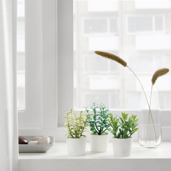 Xanela con tres plantas artificiais en pota e un vaso alto con herbas secas, traendo a natureza interior sen mantemento.