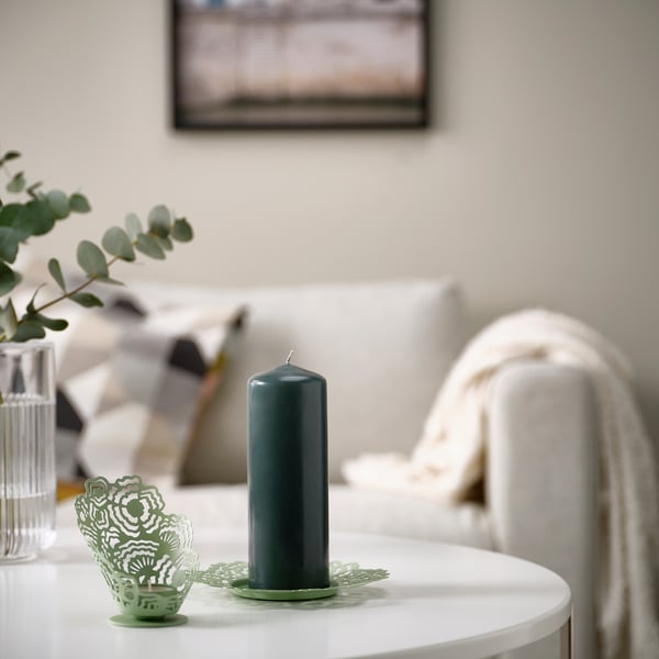 FENOMEN Vela de piar verde escuro sobre unha mesa branca. Forma cilíndrica alta e única. Colocado preto de pequenos elementos verdes decorativos, creando unha atmosfera acolledora.