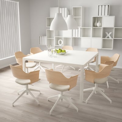 Comedor moderno con mesa branca, cadeiras xiratorias, cunca de froitas e lámpadas. Estantes con x e o. Espazo luminoso e minimalista.