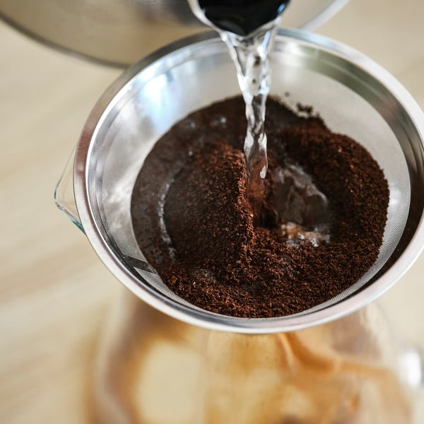 A imaxe mostra un colador metálico cheo de pos de café escuro. Está a verter auga nel, probablemente para a elaboración.
