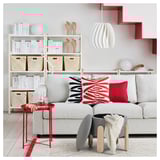 Sala de estar elegante con sofá gris en forma de L, almofadas de cores, mesa de lado vermella, estante con libros, caixas tecidas, luz colgante e elementos decorativos.