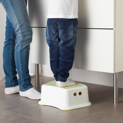 O neno está de pé sobre o taburete branco FÖRSIKTIG, axudándolles a alcanzar superficies máis altas. O taburete ten unha parte superior antideslizante e unha base estable.