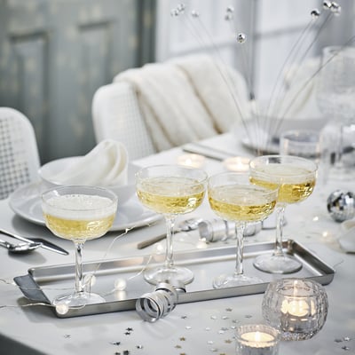 Conxunto de mesa con coupés de champaña FRÖJDA transparente, bandexa de prata, confeti estrela, fondo azul claro, sofá branco, configuración elegante.