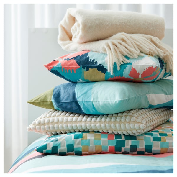 Almofadas de cores vibrantes apiladas na cama, patróns e texturas variadas, principalmente azul.