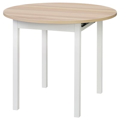 Representa unha mesa de comedor de madeira redonda e extensible con patas brancas. Está deseñado para 2-4 persoas e ten unha parte superior lisa, de cor clara.