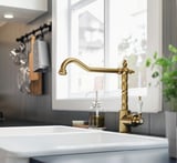 Billa de cociña dourada GLITTRAN con foca curva, con deseño de latón decorado, colocado contra un lavabo e unha xanela limpas.