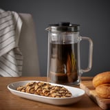 Prensa de café limpa sobre a mesa de madeira, xunto a ela un prato de galletas de chocolate e donuts azucarados.