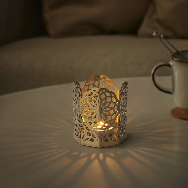 Soporte de luz de té GOKVÄLLÅ en prata, forma hexagonal, deseño floral, pequeno tamaño na mesa xunto á cunca.