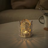 Soporte de luz de té GOKVÄLLÅ en prata, forma hexagonal, deseño floral, pequeno tamaño na mesa xunto á cunca.