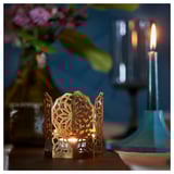 Portavelas metálico dourado con recortes florais, vela acendida dentro. Nunha mesa, xunto a unha vela azul. Decoración decorativa do fogar con luz brillante suave.