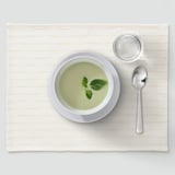 Cunca de sopa verde con culler e vaso pequeno sobre alfombra beige.